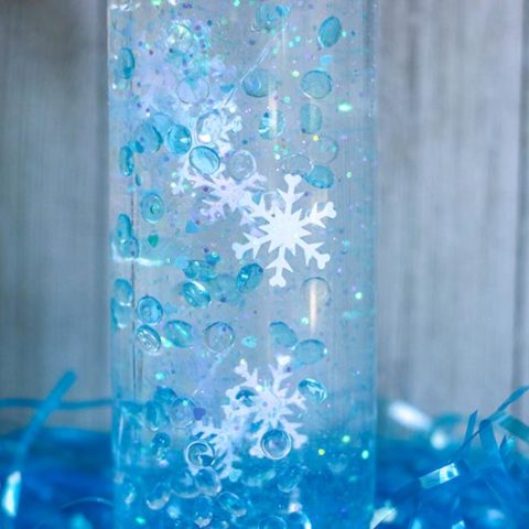 Close up of snowflake confetti, blue fish bowl beads, and glitter slowing descending in plastic bottle.