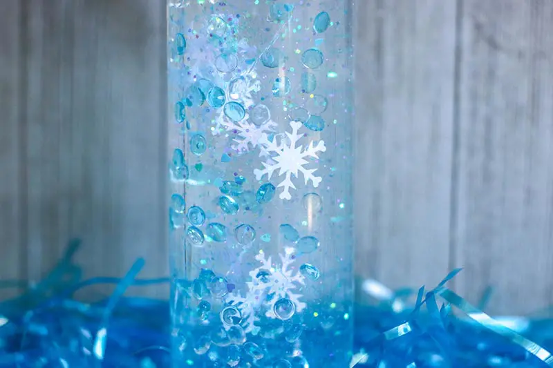 Close up of snowflake confetti, blue fish bowl beads, and glitter slowing descending in plastic bottle.