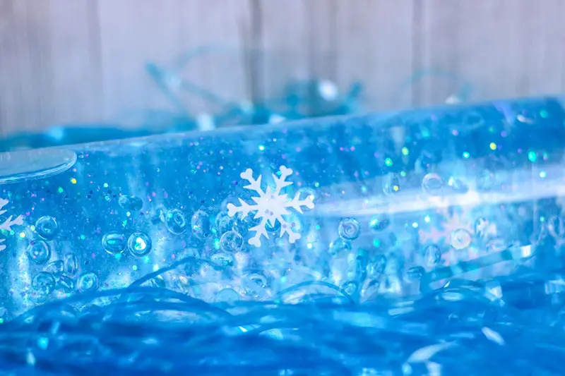 Close up of snowflake confetti suspended in glue inside bottle. Bottle laying on its side on top of blue tinsel.