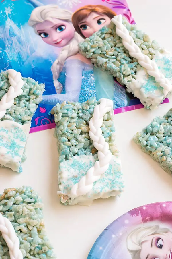Blue rice crispy treats decorated with white candy melts, blue sprinkles and braided white fondant on table with Disney Frozen napkins.