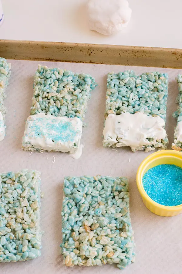 Decorating blue rice crispy treats with white candy melts and blue sugar sprinkles on baking sheet.