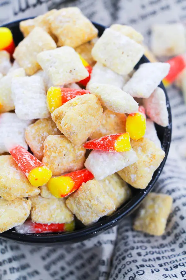 Homemade candy corn puppy chow mix in black bowl on towel that looks like book pages.