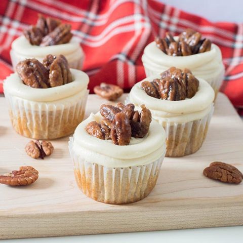 Frosted pecan pie cupcakes on a wood block with pecans scattered around and red plaid flannel cloth.