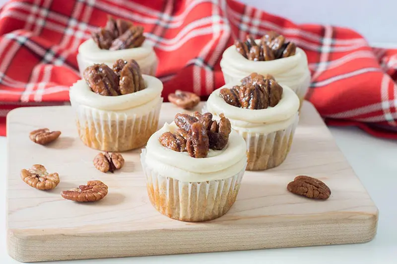 Frosted pecan pie cupcakes on a wood block with pecans scattered around and red plaid flannel cloth.