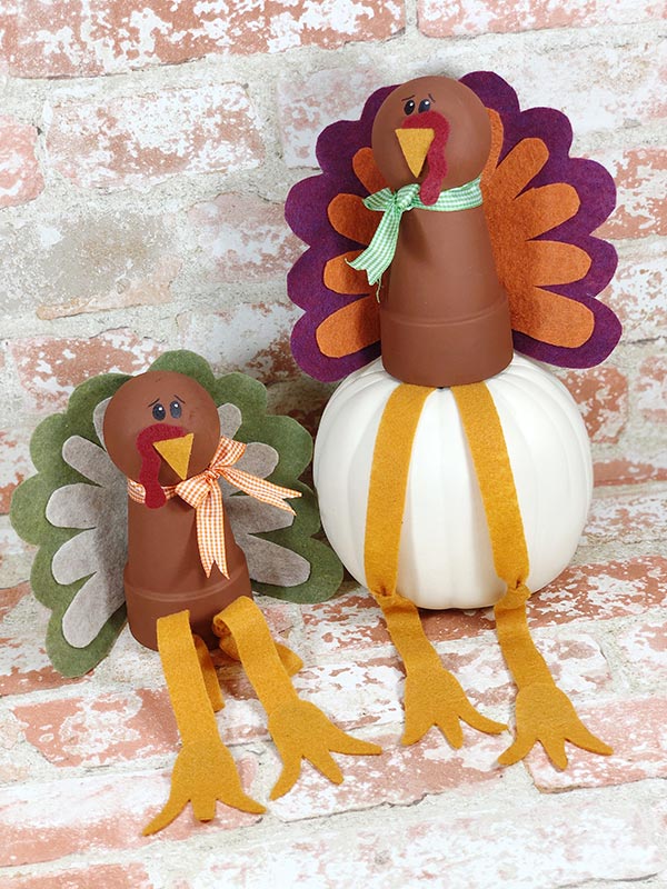 Two turkeys made out of flower pots, round wood knobs, and felt. One sitting on top of a white pumpkin and one sitting on a brick background.
