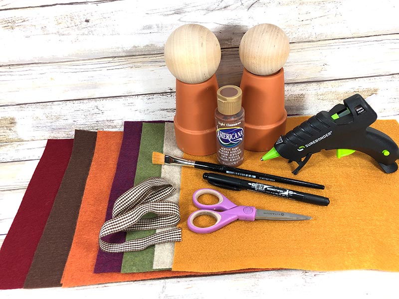 Craft felt in assorted colors, gingham ribbon, clay flower pots, wooden knobs, acrylic paint, glue gun, paint brush, marker, and scissors on a white wood background.