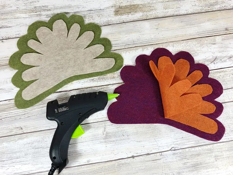 Using hot glue gun to glue craft felt together to make turkey feathers. One set is dark green and off white and the other is purple and orange.