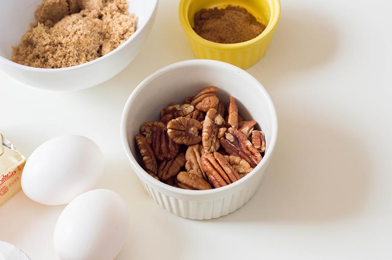Brown sugar, cinnamon, and pecans in small bowls. Eggs and butter on table next to bowls.