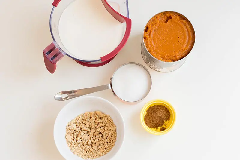 Heavy cream and sugar in measuring cups, open can of pumpkin puree, spices in small yellow bowl, and crushed graham crackers in white bowl.