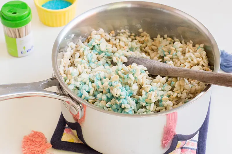Stirring rice krispies cereal into blue marshmallow mixture in silver saucepan.