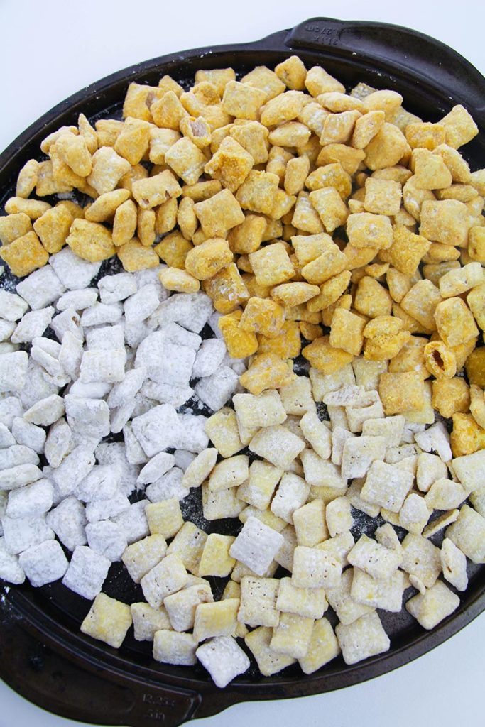 Chex cereal coated in white, yellow, and orange chocolate on a black tray.