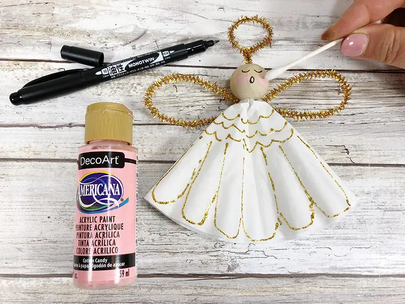 Bottle of pink craft paint and black marker laying next to angel ornament craft. Using cotton swab to apply paint to angel's face.