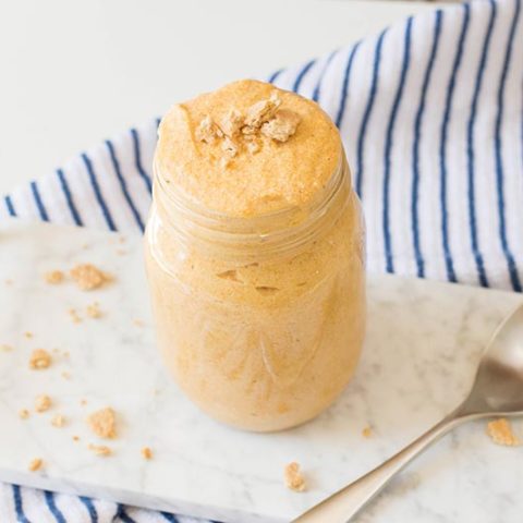 Pumpkin mousse topped with pieces of graham cracker in a glass mason jar. Jar is placed next to a spoon on a white marble serving tray with golden handles. Tray is on a white and blue striped cloth.