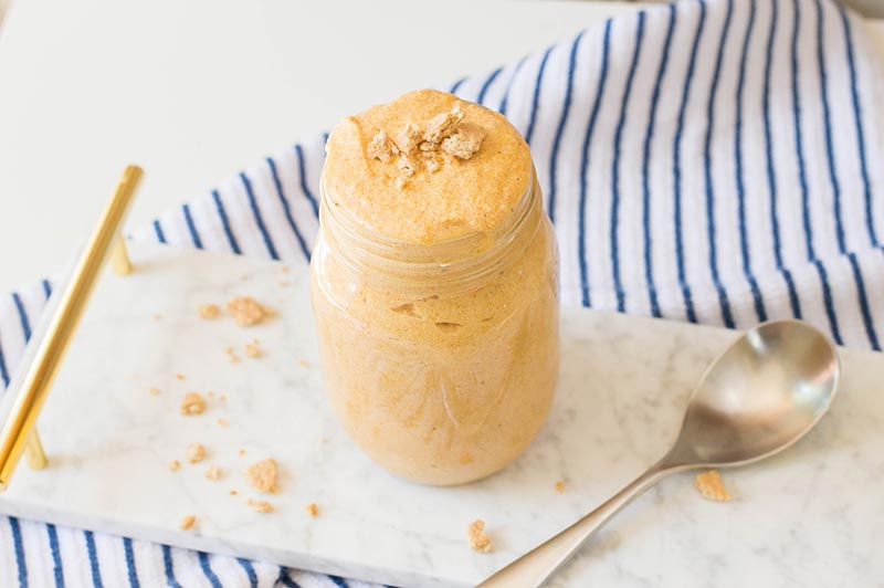 Pumpkin mousse topped with pieces of graham cracker in a glass mason jar. Jar is placed next to a spoon on a white marble serving tray with golden handles. Tray is on a white and blue striped cloth.