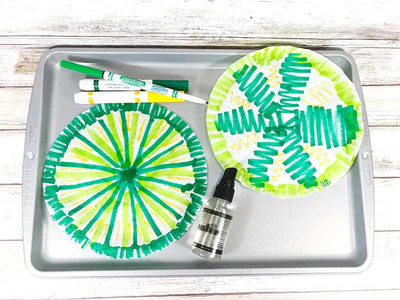 Two coffee filters colored on with green and yellow markers laying on cookie sheet.