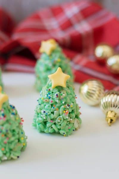 Rice crispy treats that look like Christmas trees standing upright on a white counter surrounded by small gold ornaments and a red plaid cloth.