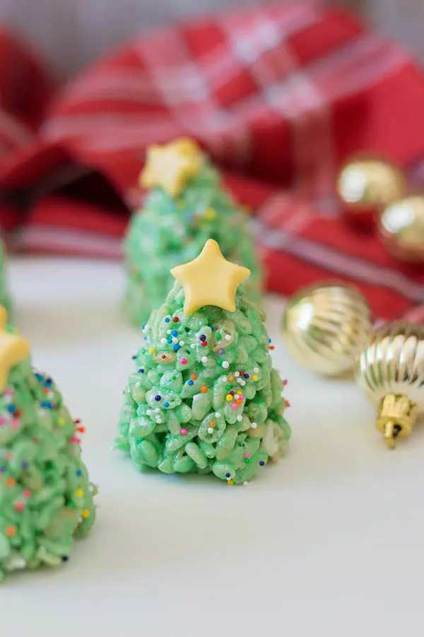 Rice crispy treats that look like Christmas trees standing upright on a white counter surrounded by small gold ornaments and a red plaid cloth.