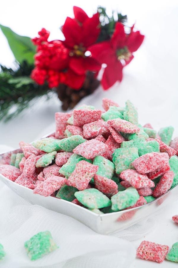 Red and green puppy chow treat made with Chex cereal in silver bowl on white table with red flowers laying behind it.