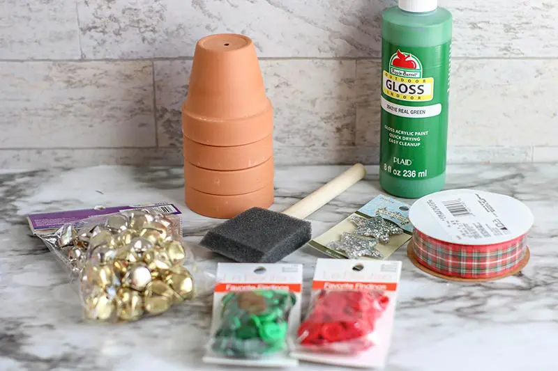 Bag of jingle bells, stack of small clay flower pots, foam paint brush, red and green buttons, green paint, and plaid ribbon for craft project.