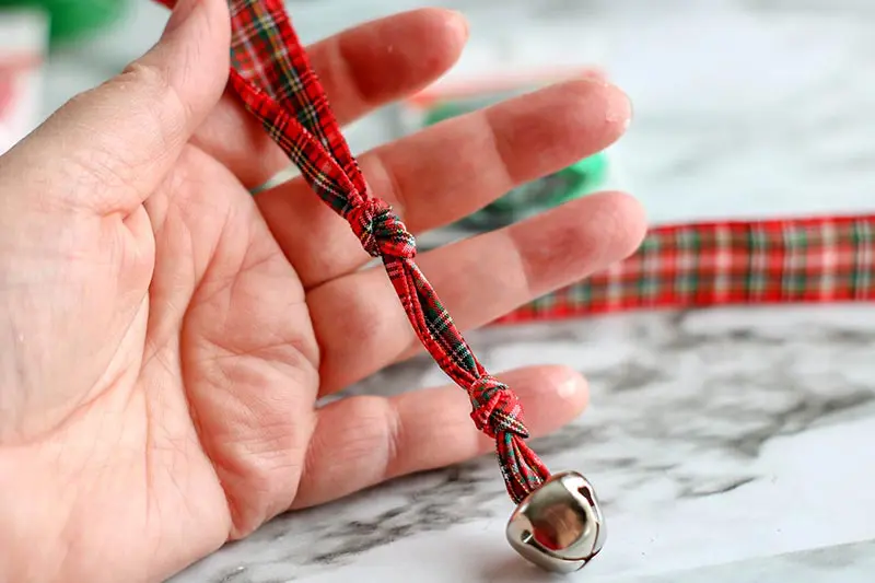 Hand holding length of red and green plaid ribbon strung through bell and knotted in two places.