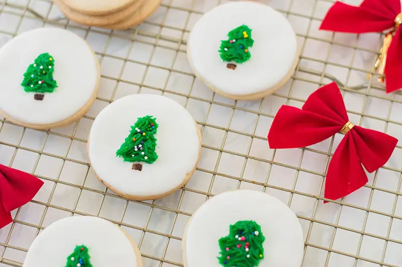 Closer view of finished sugar cookies decorated with fondant for white background with green frosting Christmas trees in the center. Cookies are on a wire rack with red bows laying around them.