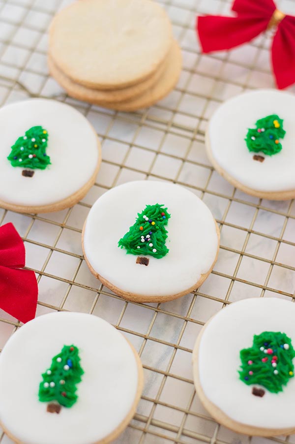 Stack of undecorated round sugar cookies on wire rack next to cookies decorated with white fondant and little Christmas trees in the center.