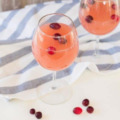 Two wine glasses with the cranberry orange drink and frozen cranberries on a white counter next to a white and blue striped cloth.