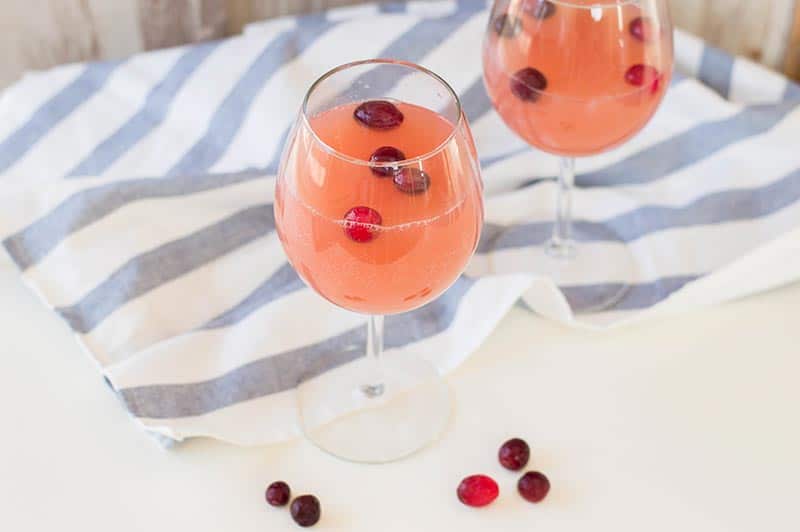 Two wine glasses with the cranberry orange drink and frozen cranberries on a white counter next to a white and blue striped cloth.