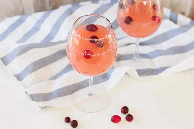 Two wine glasses with the cranberry orange drink and frozen cranberries on a white counter next to a white and blue striped cloth.