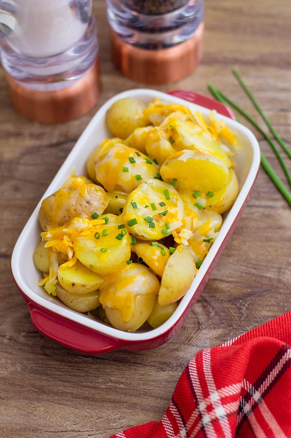 Cooked cheesy potatoes garnished with chives in small rectangular serving dish. Salt and pepper shakers and red plaid cloth on table next to dish.