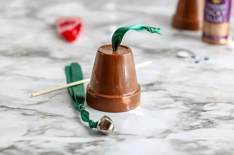 Length of green ribbon with bell on one end laying next to brown painted clay pot turned upside down with green ribbon coming out the hole.