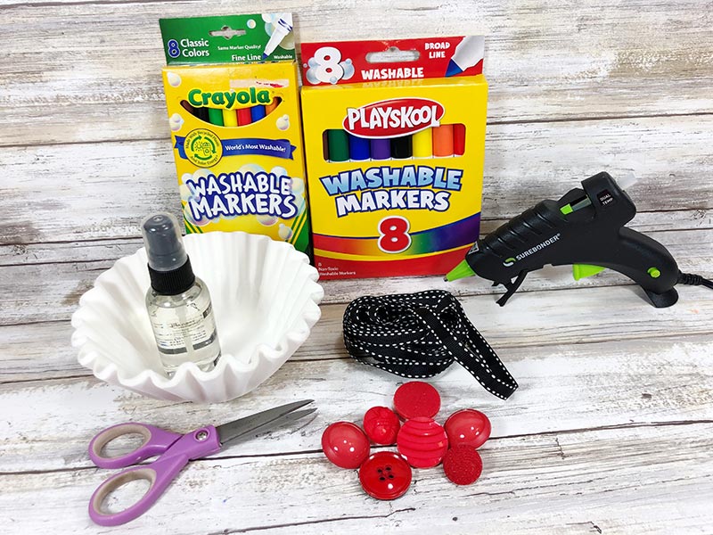 Markers, coffee filters, glue gun, ribbon, red buttons, and scissors set out on a white wood background.