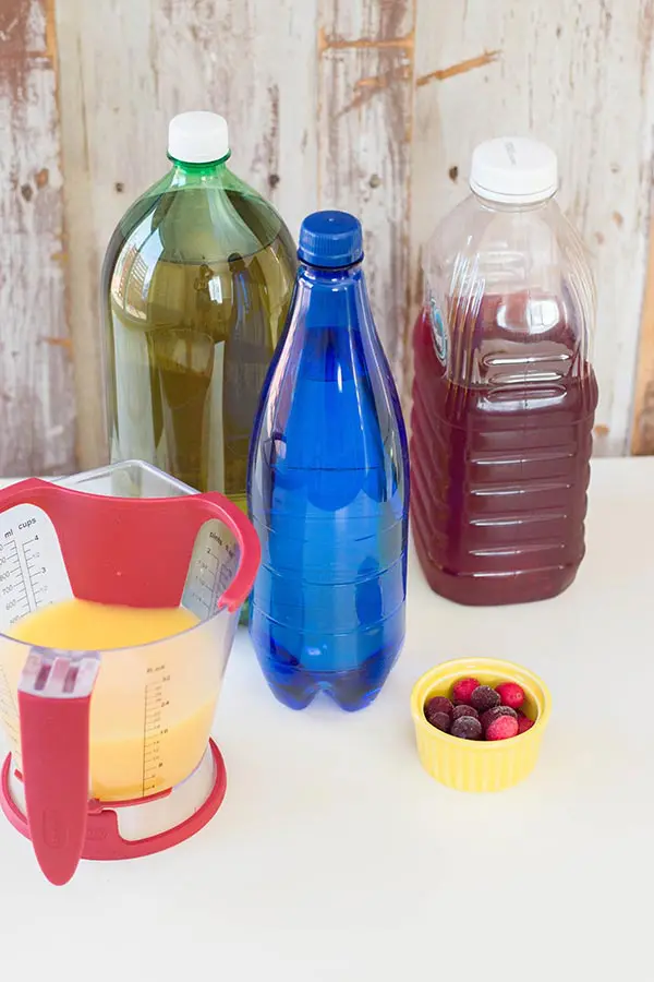 Measuring cup with orange juice, ginger ale bottle, blue bottle of sparkling water, clear plastic bottle of cranberry juice, and small yellow bowl with frozen cranberries on a white counter. Bottles have labels removed.