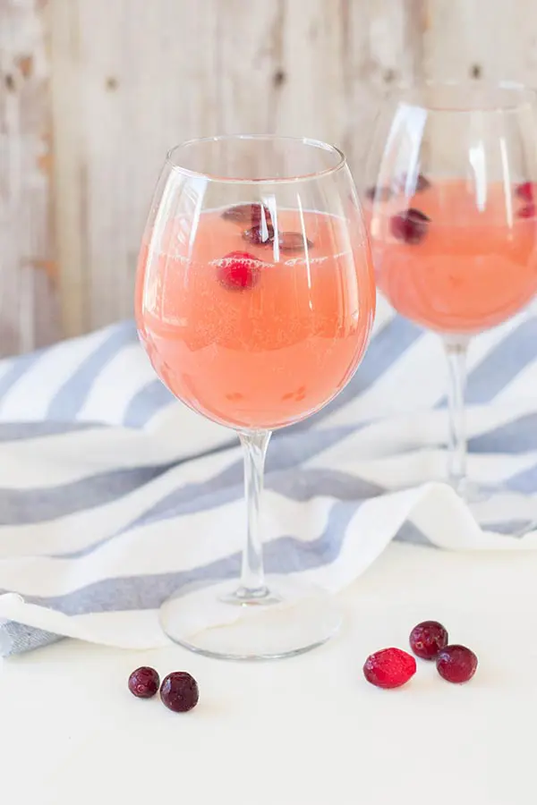Cranberry orange drink with frozen cranberries in two wine glasses with white and blue kitchen towel behind them.