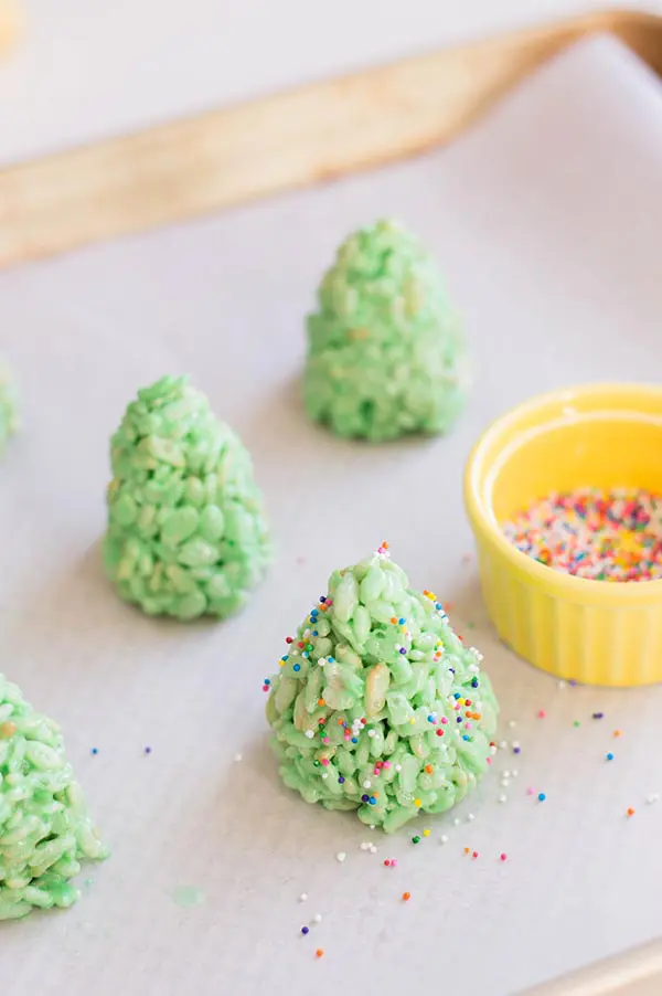 Green rice crispy treats shaped like trees standing on wax paper. Small yellow ramekin with rainbow nonpareils sprinkles in it and some sprinkled over the treats.