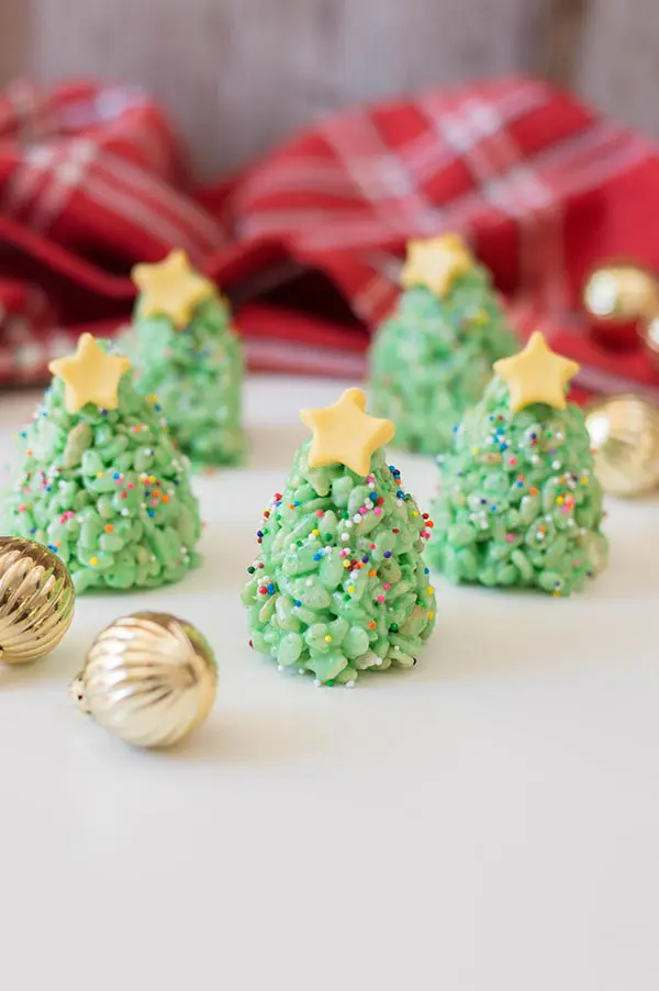 Green rice crispy treats shaped and decorated like Christmas trees with rainbow sprinkles and yellow fondant star on top. Treats are standing on a white countertop with small round gold ornaments and red plaid cloth around them.