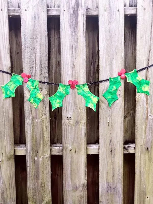 Ribbon banner with three coffee filter holly leaves and berries hanging along a wooden fence.
