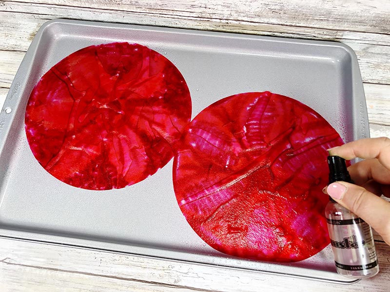 Two red colored coffee filters on cookie sheet and hand spraying water on them with small bottle.