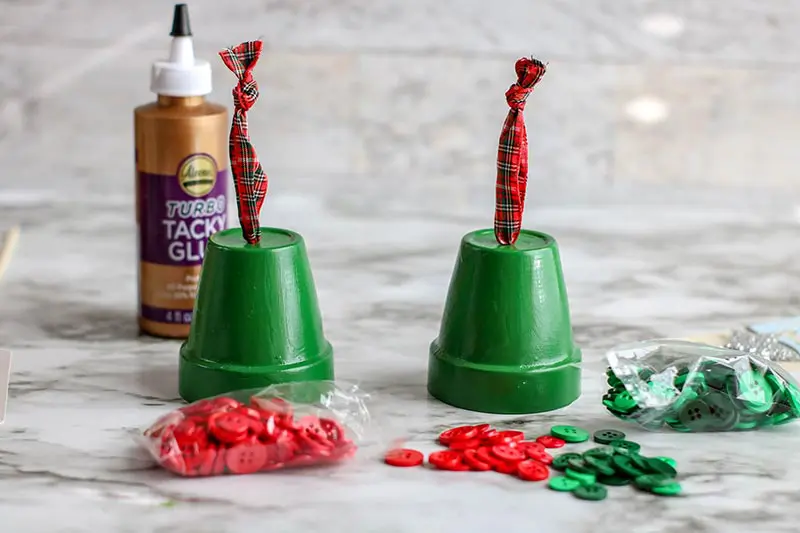 Two green painted clay pots set upside down with plaid ribbon coming out of hole. Red and green buttons and craft glue next to them on the table.