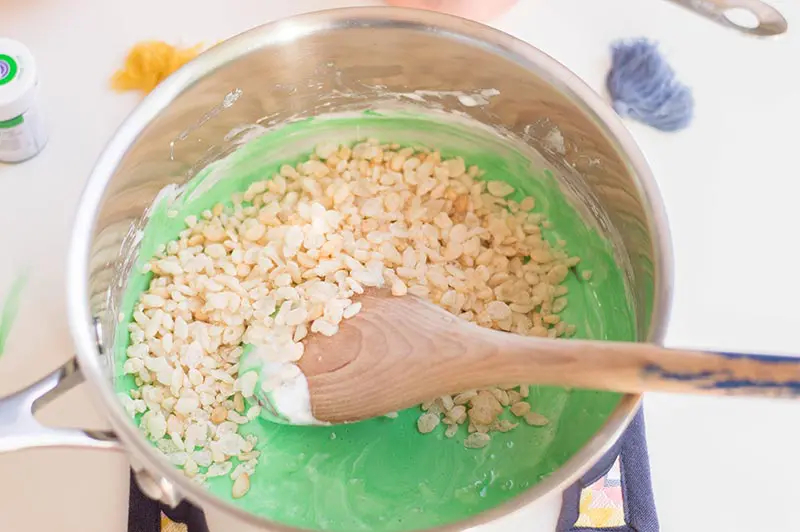 Stirring rice crispy cereal with wooden spoon into green tinted melted marshmallows in sauce pan.