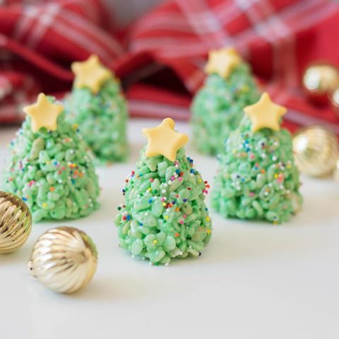 Five completed Christmas tree rice crispy treats with yellow fondant stars on top. Trees are standing up on a white counter with a plaid cloth in back and small golden ornaments scattered around.