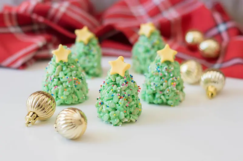 Five completed Christmas tree rice crispy treats with yellow fondant stars on top. Trees are standing up on a white counter with a plaid cloth in back and small golden ornaments scattered around.