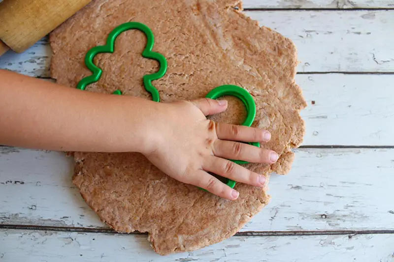 Light brown playdough rolled out with rolling pin and kid hand pressing green plastic gingerbread men shaped cookie cutters into it.