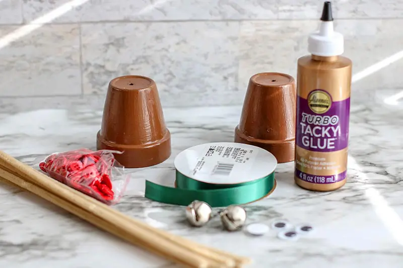 Clay flower pots, green ribbon, red buttons, jingle bells, light brown pipe cleaners, googly eyes, and craft glue sitting on marble counter top.