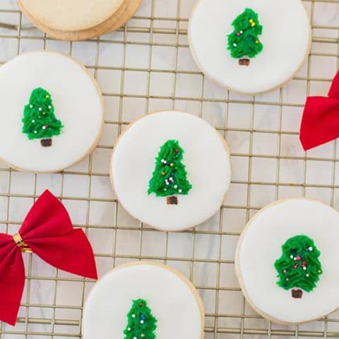 Five circle sugar cookies decorated white with tree in the center. Cookies are laying out on a wire rack with red bows laying nearby.