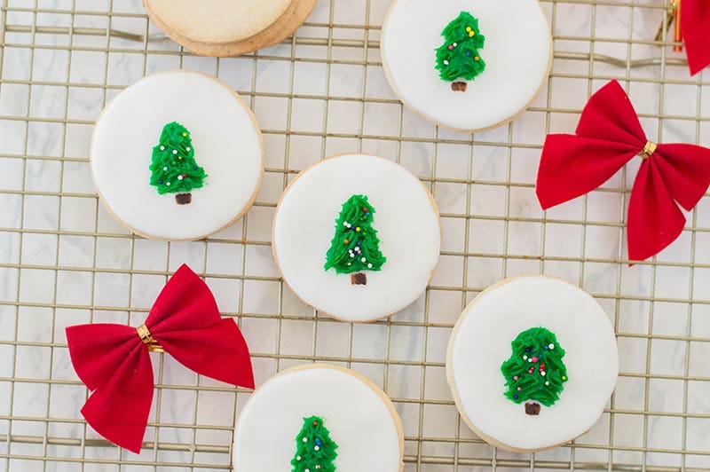 Five circle sugar cookies decorated white with tree in the center. Cookies are laying out on a wire rack with red bows laying nearby.