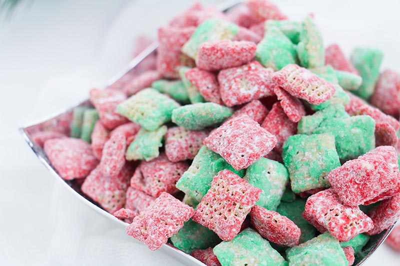 Close view of red and green Christmas puppy chow mix in square silver bowl on white table.