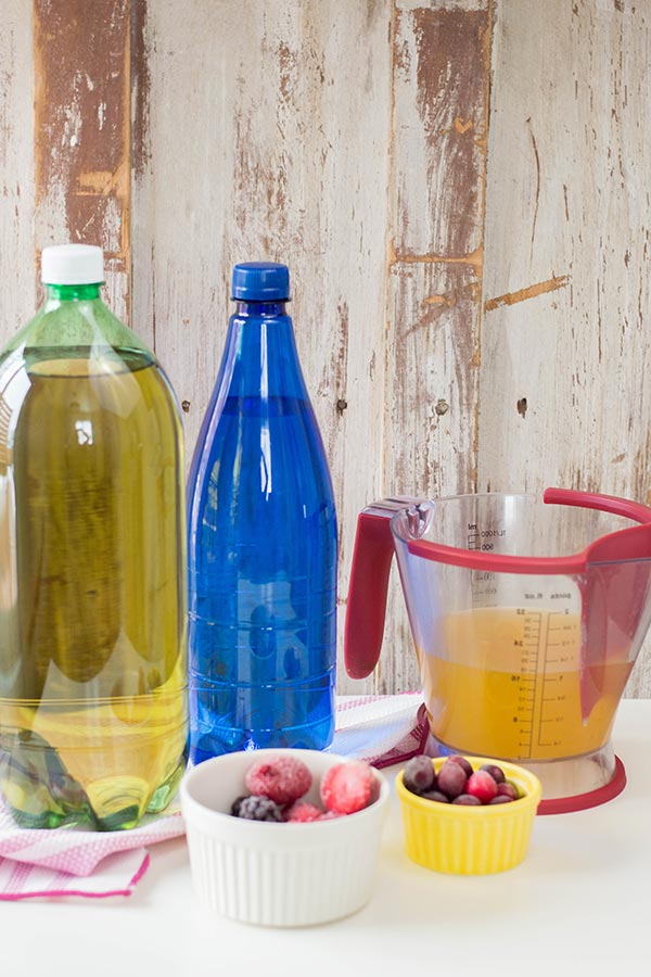 Bottle of ginger ale, blue bottle of sparkling water, measuring cup with pineapple juice, and small bowls with frozen fruit on a white counter top.