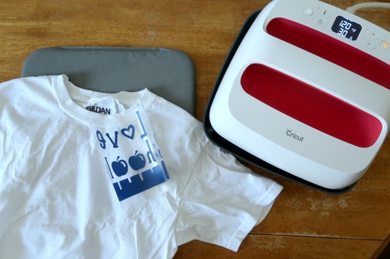 Blank white t shirt laying on gray mat on table with Cricut EasyPress 2 and iron on design.