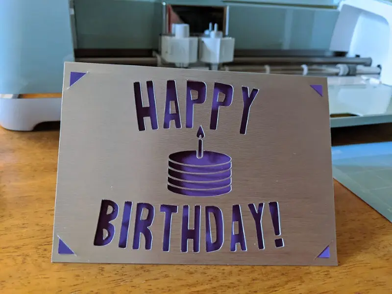 Completed silver and purple birthday card standing on table in front of Cricut machine.
